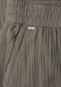 Gros plan sur la ceinture élastique d’un pantalon gris texturé avec poche latérale et petite étiquette métallique de la marque « Street One » attachée près de la taille.