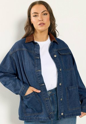 Kvinde iført stor, mørkeblå denimjakke med brun krave, hvid skjorte og matchende blå jeans, stående op ad en ensfarvet væg.