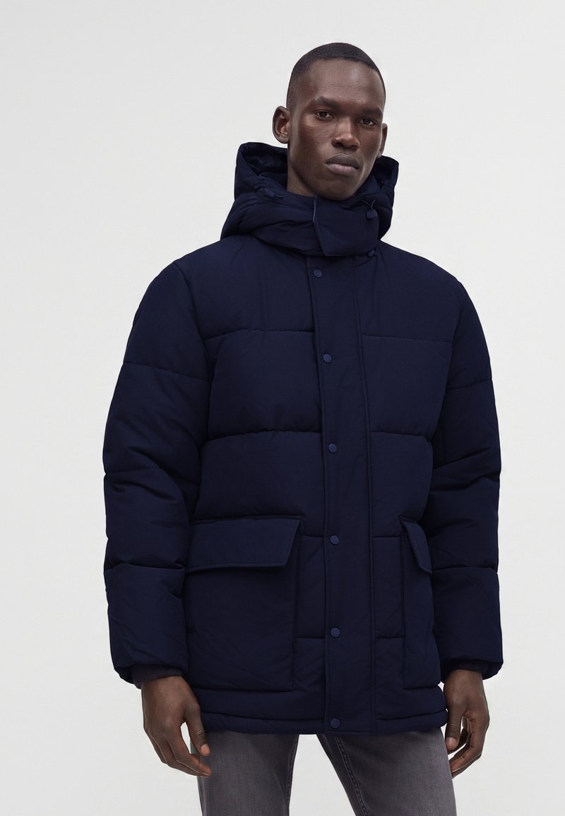 Homme portant une veste d'hiver matelassée bleu marine avec de grandes poches avant et une capuche, debout devant un fond clair uni.