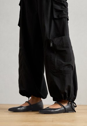 Pieds portant des ballerines Mary Jane noires à bout pointu avec des brides cloutées, associées à un pantalon cargo noir ample resserré aux chevilles avec des liens.