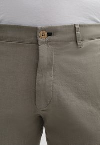 I pantaloni beige con texture presentano una chiusura a bottone, un sottile motivo intrecciato e passanti per cintura. Il tessuto è liscio con una leggera lucentezza.