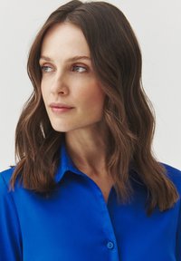Blue collared shirt in smooth fabric with buttons, featuring a sleek design and a close fit, complemented by long, wavy brown hair.