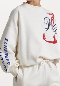 Vit sweatshirth med hög krage, långa ärmar, blå text som säger "KVALITETSPLAGG" på ärmen, och en röd ankargrafik på framsidan.