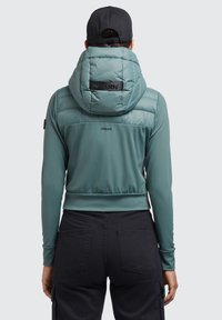 Jachetă puffer teal cu glugă mare, material elastic și siluetă ajustată. Prezintă un logo pe spate și tiv elastic. Este purtată cu pantaloni negri.