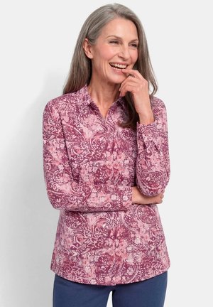 Middle-aged woman with gray hair smiles, wearing a long-sleeve pink floral patterned shirt and blue pants against a neutral background.
