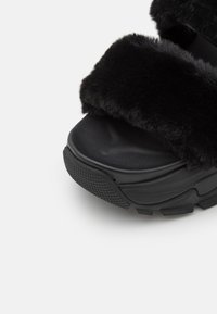 Kraftig sort sandal med tyk tekstureret sål og brede, bløde pelsremme over tæerne og fodområderne.