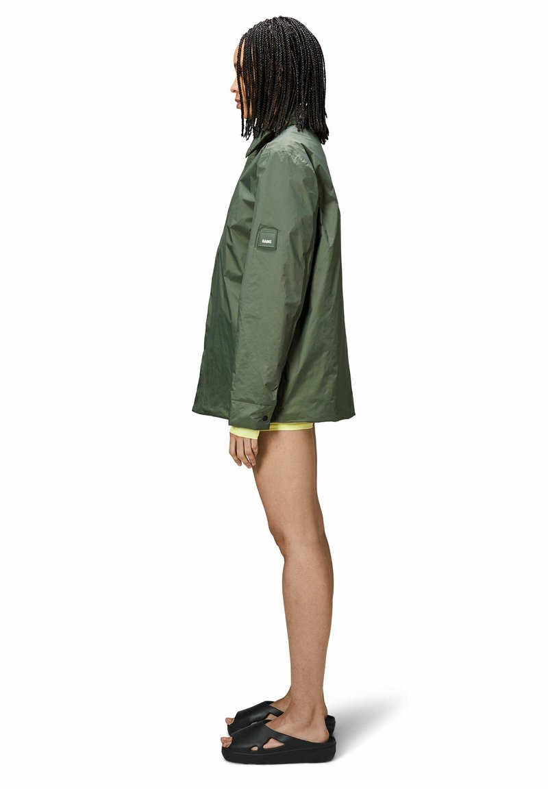 Veste imperméable vert olive à coupe ample, présentant un badge sur la manche et des accents de boutons. Portée sur un vêtement jaune, associée à des sandales noires.