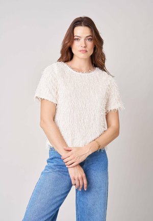 Vrouw met lang bruin haar, gekleed in een gestructureerde witte top met korte mouwen en blauwe spijkerbroek, staand met gekruiste handen voor zich.