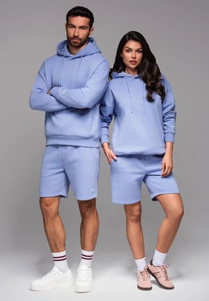 Haut à capuche bleu clair et shorts en tissu doux ; le haut à capuche possède une poche kangourou, les shorts disposent d'une taille élastique et d'un branding discret.