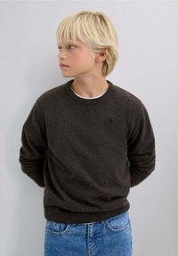 Pull en tricot gris foncé avec un col rond et des poignets côtelés, associé à un jean en denim bleu clair. Présente un petit logo bleu sur la poitrine.