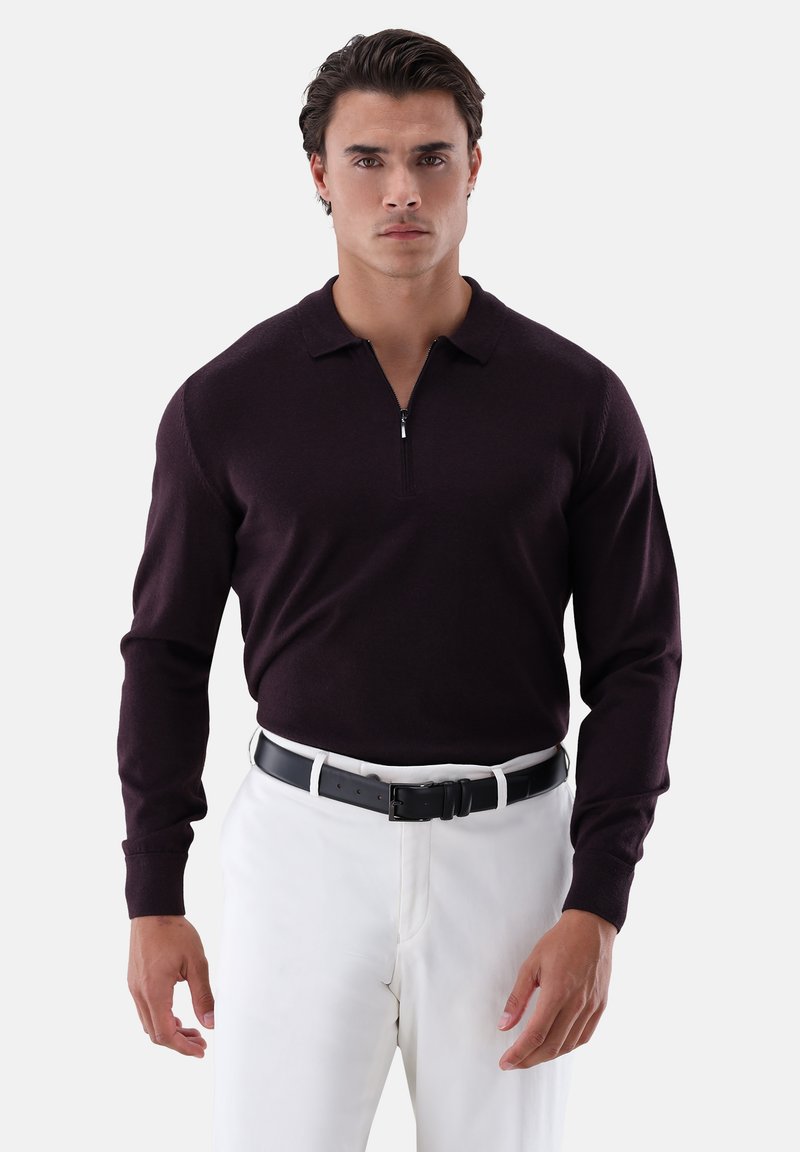 Dunkellila langärmeliger Pullover mit durchgehendem Reißverschluss und Kragen; getragen mit weißen Hosen und einem schwarzen Gürtel. Glatter Stoff und tailliertes Design.