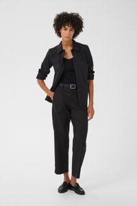 Donna con capelli ricci che indossa una camicia nera abbottonata sopra una canotta nera, pantaloni neri con cintura e mocassini neri, in piedi con una mano in tasca.