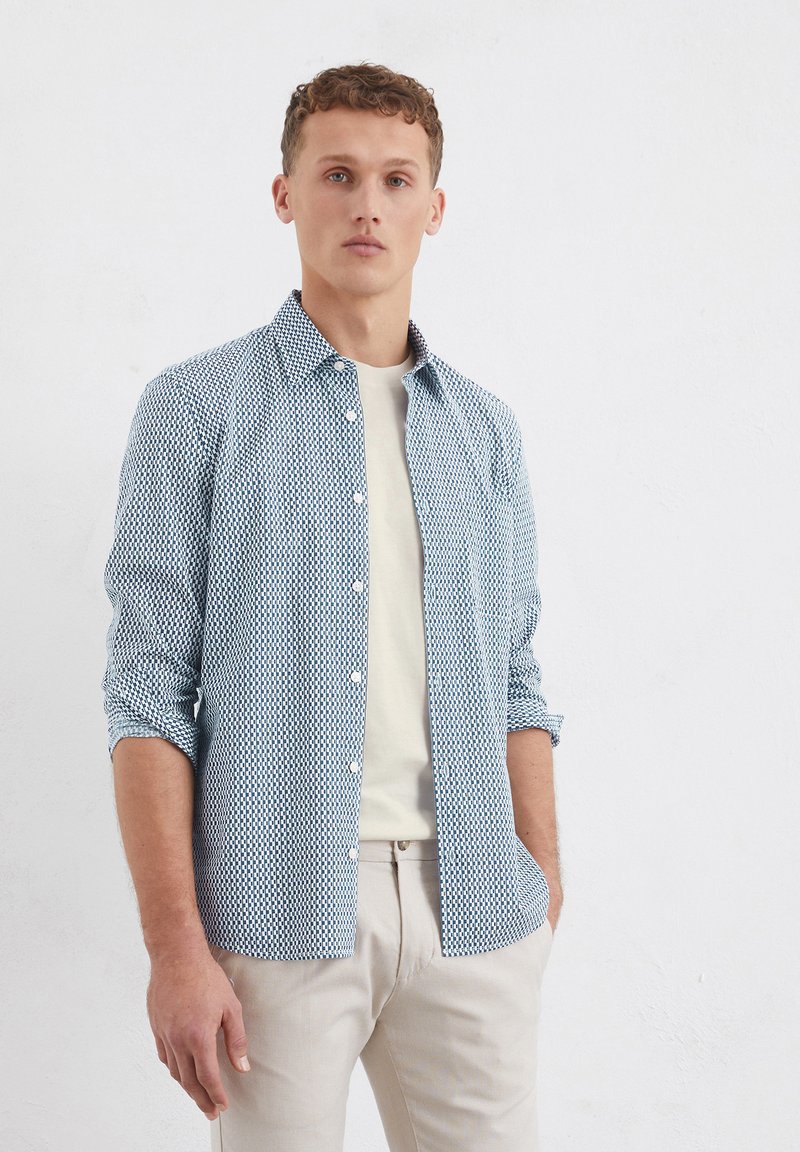 Button-up shirt in light blue with a geometric pattern. Material appears lightweight. Styled with rolled sleeves and paired with a light-colored undershirt.
