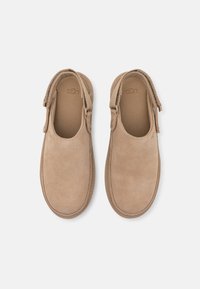Tan suede neslidinčios bateliai su apvaliais pirštais ir lygiu paviršiumi. Yra prekės ženklą turinti vidpadis ir subtili elastinga šoninė dalis, kad būtų lengva užsidėti.