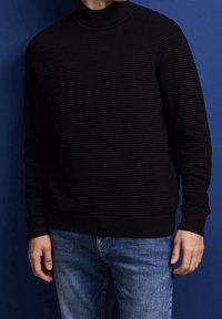 Svart pullovertröja med ribbad textur och hög krage, kombinerad med ljusblå jeans. Bakgrunden är en enfärgad mörkblå vägg.