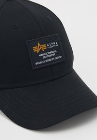 Casquette de baseball noire en tissu avec une couronne structurée. Présente une étiquette orange avec un logo et du texte, des accents cousus et un bord courbé.