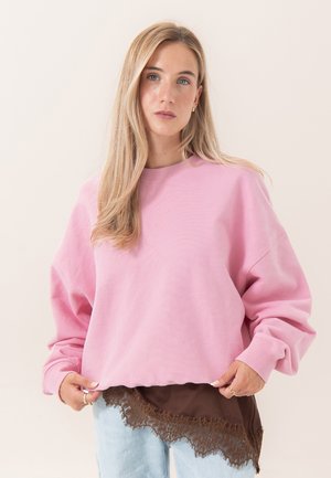 Jeune femme aux longs cheveux blonds porte un pull rose ample par-dessus un top marron orné de dentelle et un jean bleu clair, debout devant un fond uni.
