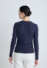 Marineblauw shirt met lange mouwen in ribbeltextuur en een ronde halslijn, gedragen met witte gewatteerde broeken; zichtbare hand en grote oorring.