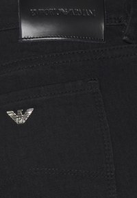 Svart denimtyg med en liten silverfärgad örnlogotyp på fickan, med en slät läderetikett präglad med "EMPORIO ARMANI" ovanför.