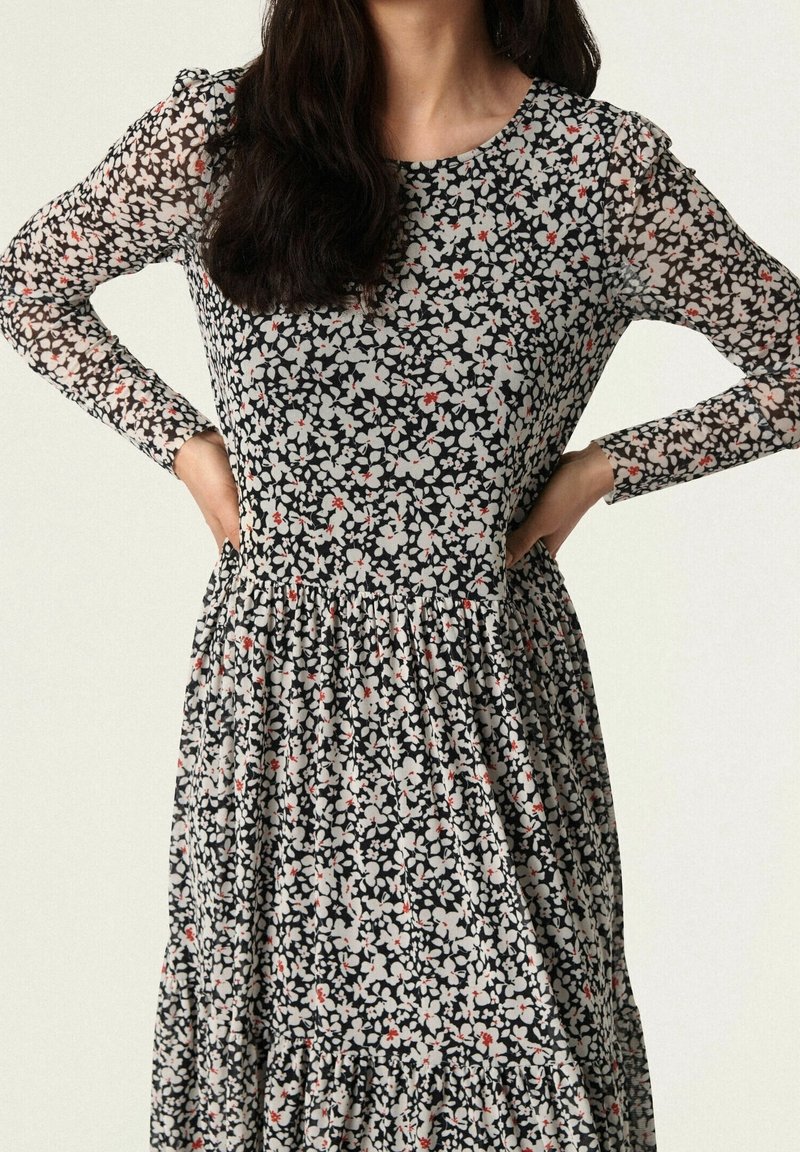 Vestido floral con base negra, con patrones de flores blancas y rojas, mangas largas transparentes, escote redondo y falda de volantes.