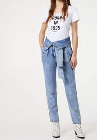 Camiseta blanca con texto negro "NACIDO EN 1995", combinada con pantalones de mezclilla azul de cintura alta con un detalle de lazo en la cintura. Botas de tobillo con estampado de estrellas.