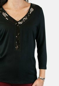 Top negro de manga larga con escote en V, que presenta un panel central de lentejuelas negras y acentos decorativos a lo largo del escote.