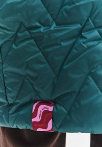 Prošívaná bunda v tmavě zeleno-modré barvě, s cikcak vzorem a látkovou štítkem s barevnými vzory a textem na spodním okraji. Měkká, lesklá textury.