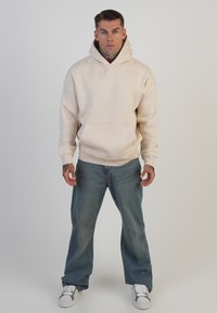 Bēša kapuce ar priekšējo kabatu un auklu, apvienota ar platajām zils džinsiem un baltiem sporta apaviem. Gluda auduma, minimāls zīmols.