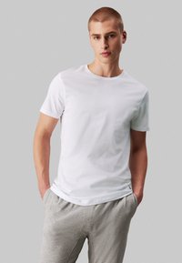 T-shirt en coton blanc à manches courtes et col rond, porté avec un pantalon à cordon gris. Design simple sans motifs ni graphiques visibles.