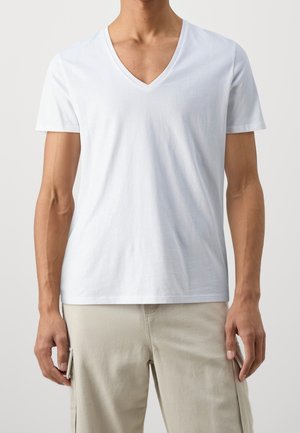 Homme portant un t-shirt blanc uni col en V à manches courtes et un pantalon beige clair avec des poches latérales, debout devant un fond uni.