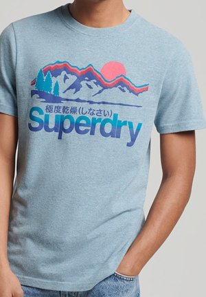 Lichtblauw t-shirt met kleurrijke bergafbeelding, roze zon, blauwe bomen en de tekst "Superdry" in het blauw met daarboven Japanse karakters.