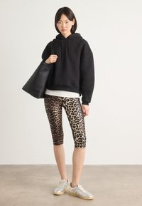 Zwarte korte hoodie, leggings met luipaardprint en zilveren sneakers; model houdt een zwarte draagtas vast; neutrale achtergrond en betonnen vloer.
