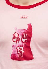 Κοντινή εικόνα ροζ πουκάμισου με κόκκινη τρέσα και κόκκινο αφηρημένο τύπωμα προσώπου κάτω από τη λέξη "Aries" στο λαιμό.