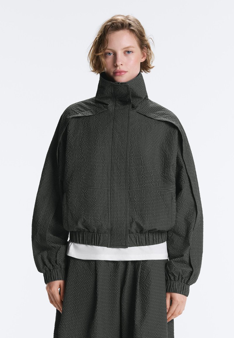 Femme portant une veste texturée gris foncé à col montant avec poignets et taille élastiques, associée à un pantalon assorti et un t-shirt blanc en dessous.