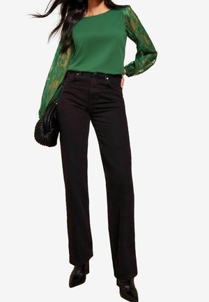Kvinde iført en grøn bluse med blonder på ærmerne, højtaljede sorte jeans, sorte spidse støvler og holder en sort quiltet håndtaske.