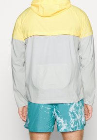 Muž v žluté a světle šedé bundě s kapucí a tyrkysových šortkách s vzorem, zobrazený zezadu na jednobarevném pozadí.