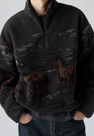 Sudadera negra de forro polar con cremallera hasta el pecho, con patrones abstractos de animales en marrón y verde, usada con las manos en los bolsillos y jeans oscuros.