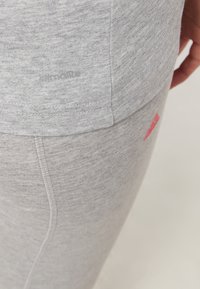 Šedé sportovní oblečení s měkkou texturou, přiléhavým střihem, zaobleným lemem a malým červeným logem na boční straně legín.