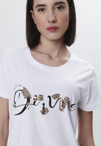 Camiseta de algodón blanca con un texto en negro tipo script que dice "Divine", adornada con detalles en tono dorado, incluyendo strass y un detalle de imperdible.