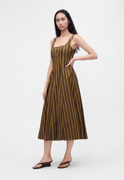 Femme debout, main sur la hanche, portant une robe midi sans manches à rayures verticales marron et noir et des sandales compensées noires.
