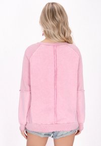 Felpa rosa chiaro con dettagli a costine, dotata di scollo a barchetta, maniche lunghe e un particolare cucito a costine sulla parte posteriore. Lunghezza corta, vestibilità rilassata.