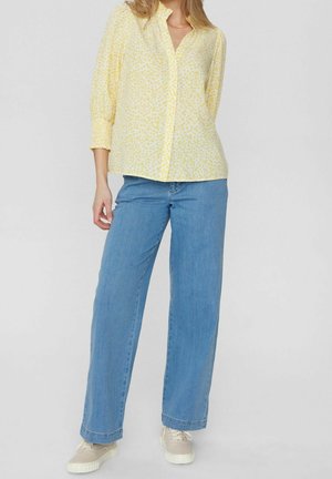 Femme portant un chemisier floral jaune clair, un jean bleu à jambes larges et des baskets beiges, debout devant un fond uni.