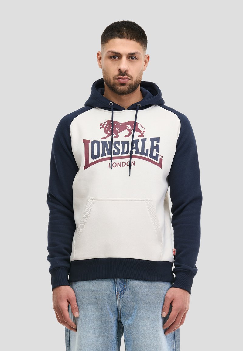 Άνδρας που φοράει φούτερ με κουκούλα Lonsdale London σε ναυτικό μπλε και λευκό με λογότυπο λιονταριού, συνδυασμένο με ανοιχτόχρωμο τζιν, στέκεται μπροστά σε απλό φόντο.