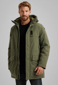 Groene parka jas met een rits en drukknoopsluiting, voorzien van een capuchon, twee voorkruiszakken en een stoffen label op de borst.