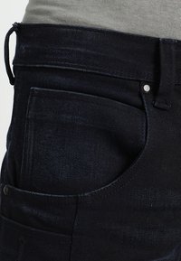Mörkblå denimjeans med en texturerad yta, med en bakficka och metallfästen. Midjebandet är utformat för en snug passform.