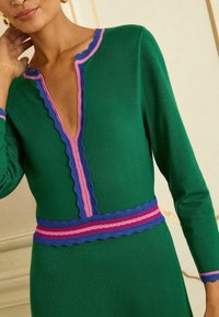 Rochie verde tricotată cu decolteu adânc în V, având margini ondulate roz și albastre de-a lungul decolteului și a taliei, cu dungi orizontale.