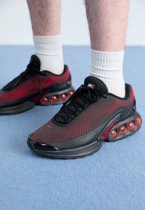 Personne portant des baskets de sport noires et rouges avec des semelles amorties à air et des chaussettes blanches, debout sur un sol recouvert de moquette bleue.