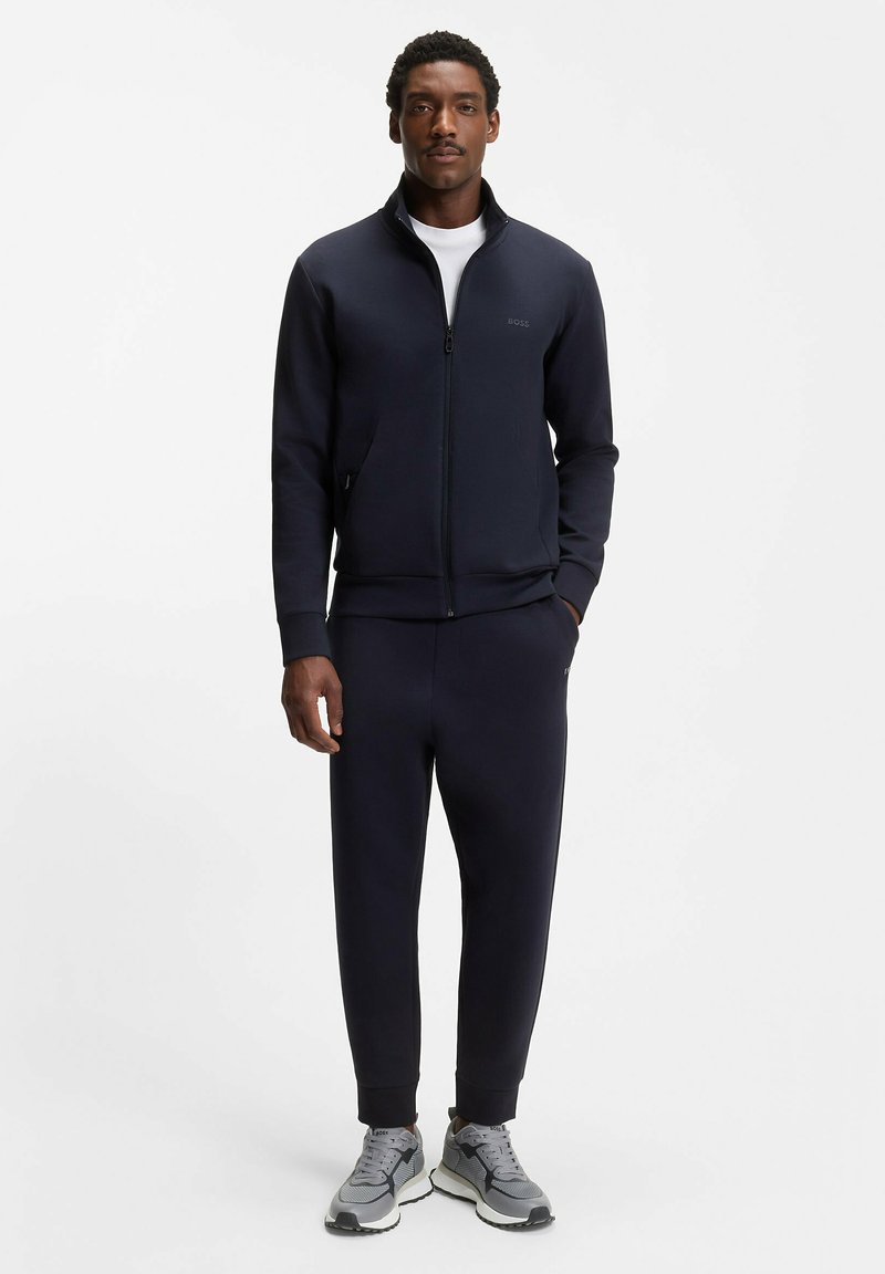 Mężczyzna ubrany w granatową rozpinaną bluzę, pasujące dresy, białą koszulę i szare tenisówki, stojący na tle jednolitego białego tła.