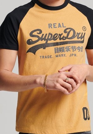 Mężczyzna noszący musztardowo-żółtą i czarną koszulkę SuperDry z japońskimi znakami, złotą bransoletkę i pierścionek na palcach.