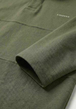 Olivengrønn langermet skjorte med synlige sømmer og liten hvit tekst «CISZBEE» nær skulderområdet.